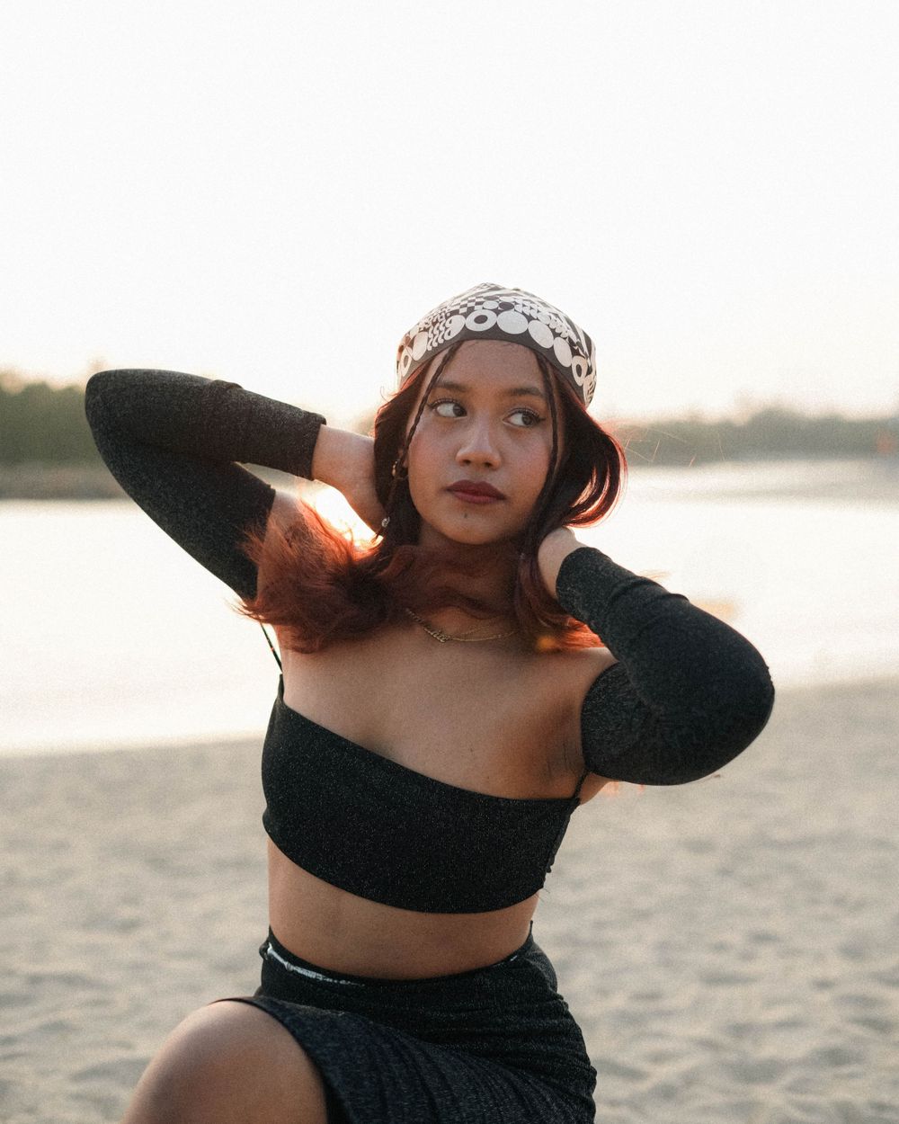Sunset beach portrait with black crop top, matching skirt and patterned bandana against water backdrop.