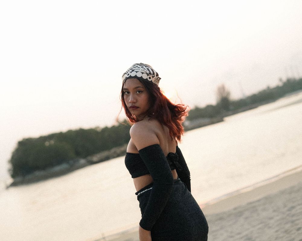 Dynamic beach pose with black two-piece outfit and patterned headwear against waterfront view.