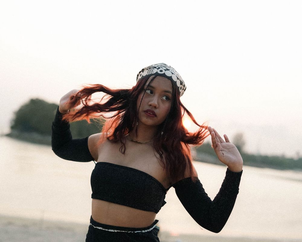Playful hair flip portrait in black two-piece outfit with patterned bandana near water.