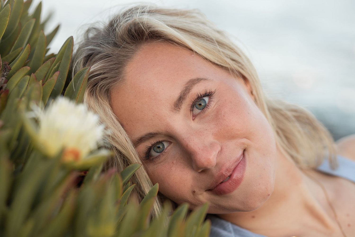 Close up of blonde woman with blue eyes smiling leaning head against blurred leaves on Maui