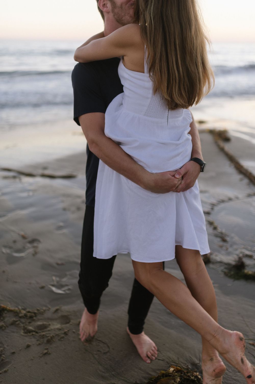 A couple embraces barefoot on the beach at sunset with waves in the background.