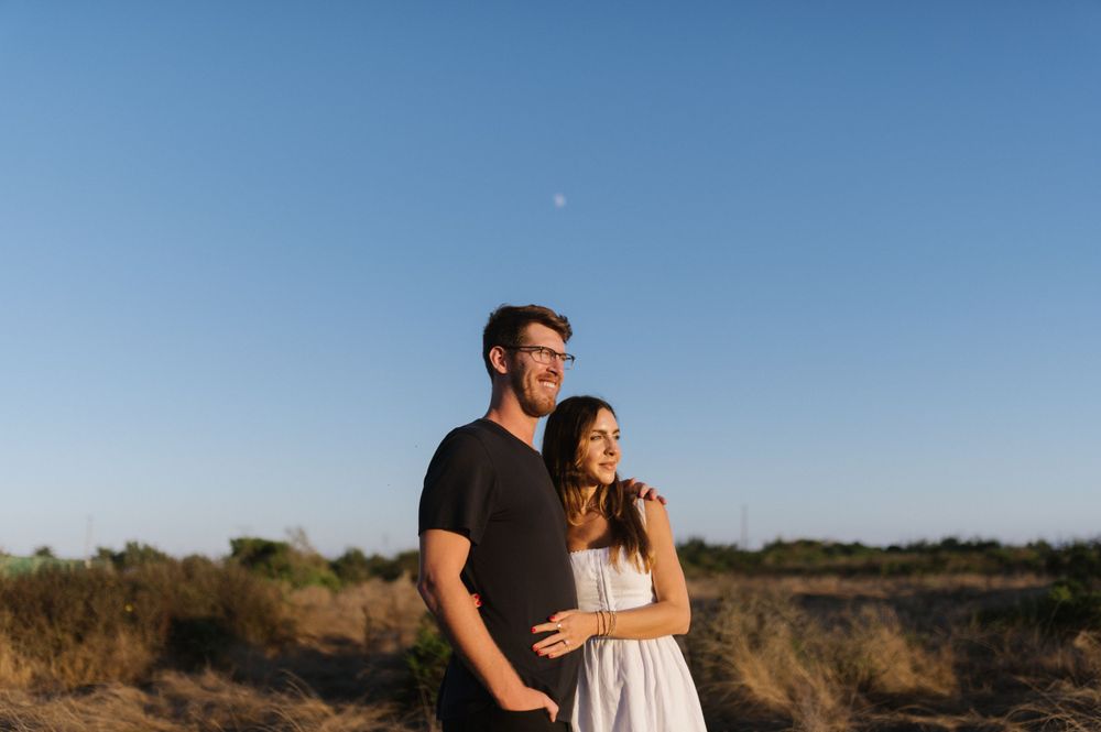 A couple in coordinated black and white outfits poses together against a blue sky and coastal landscape.