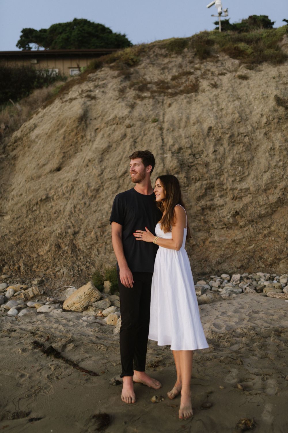 A couple embraces on a sandy beach against a rocky cliff backdrop during golden hour.