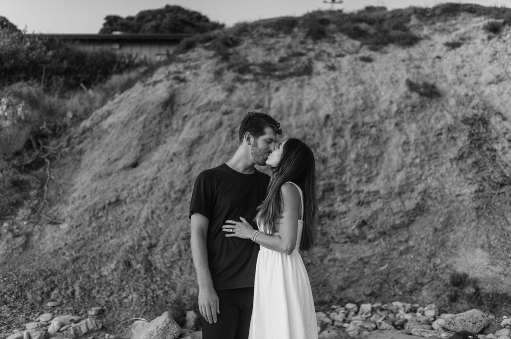 A romantic black and white portrait of a couple embracing against a hill backdrop during a photoshoot.
