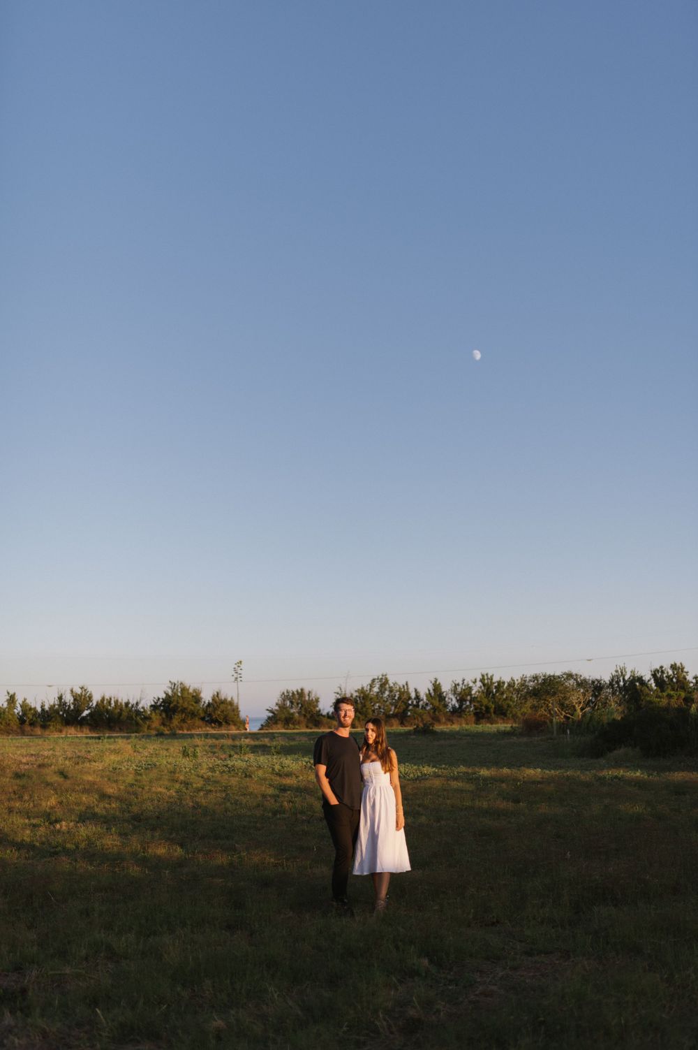 A couple stands in a grassy field under a clear sky with a visible moon.