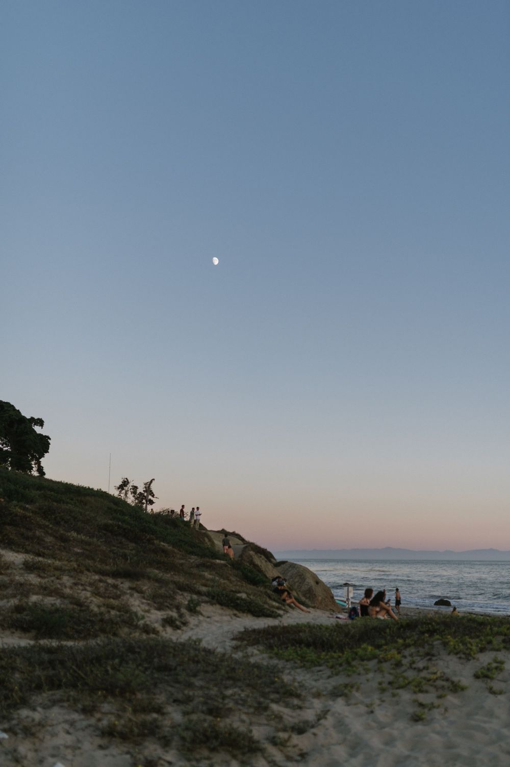 Scenic coastal landscape at dusk with moon visible in blue sky.