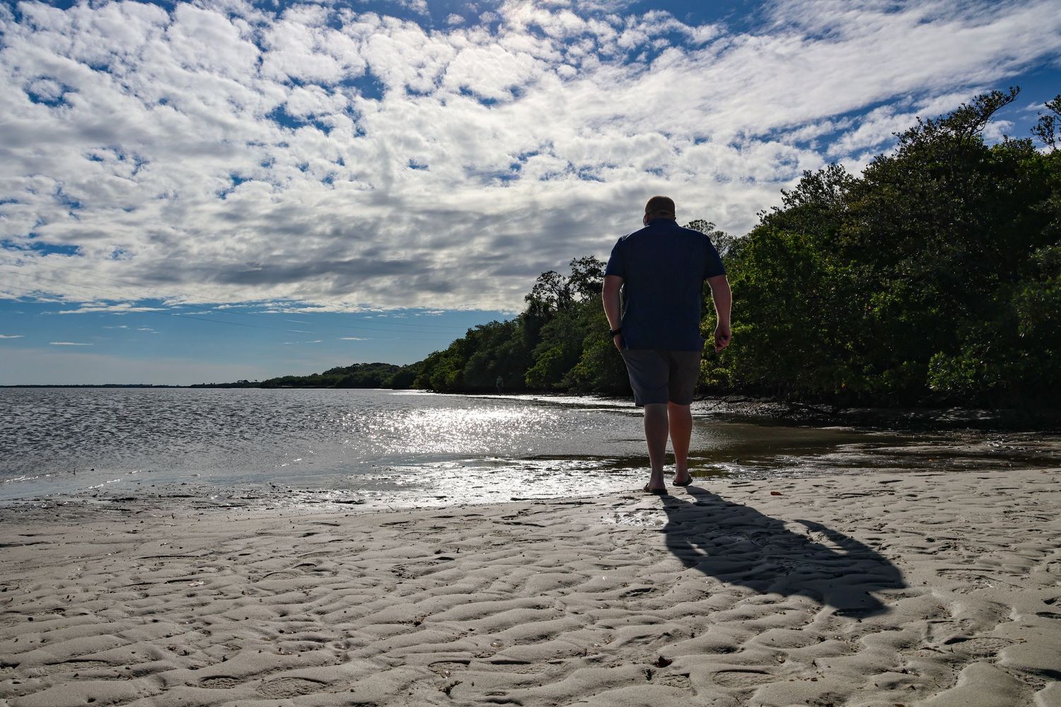 A person walks on Phillipe Park's pristine beach with scattered clouds in the sky while trees line the shoreline in the distance.