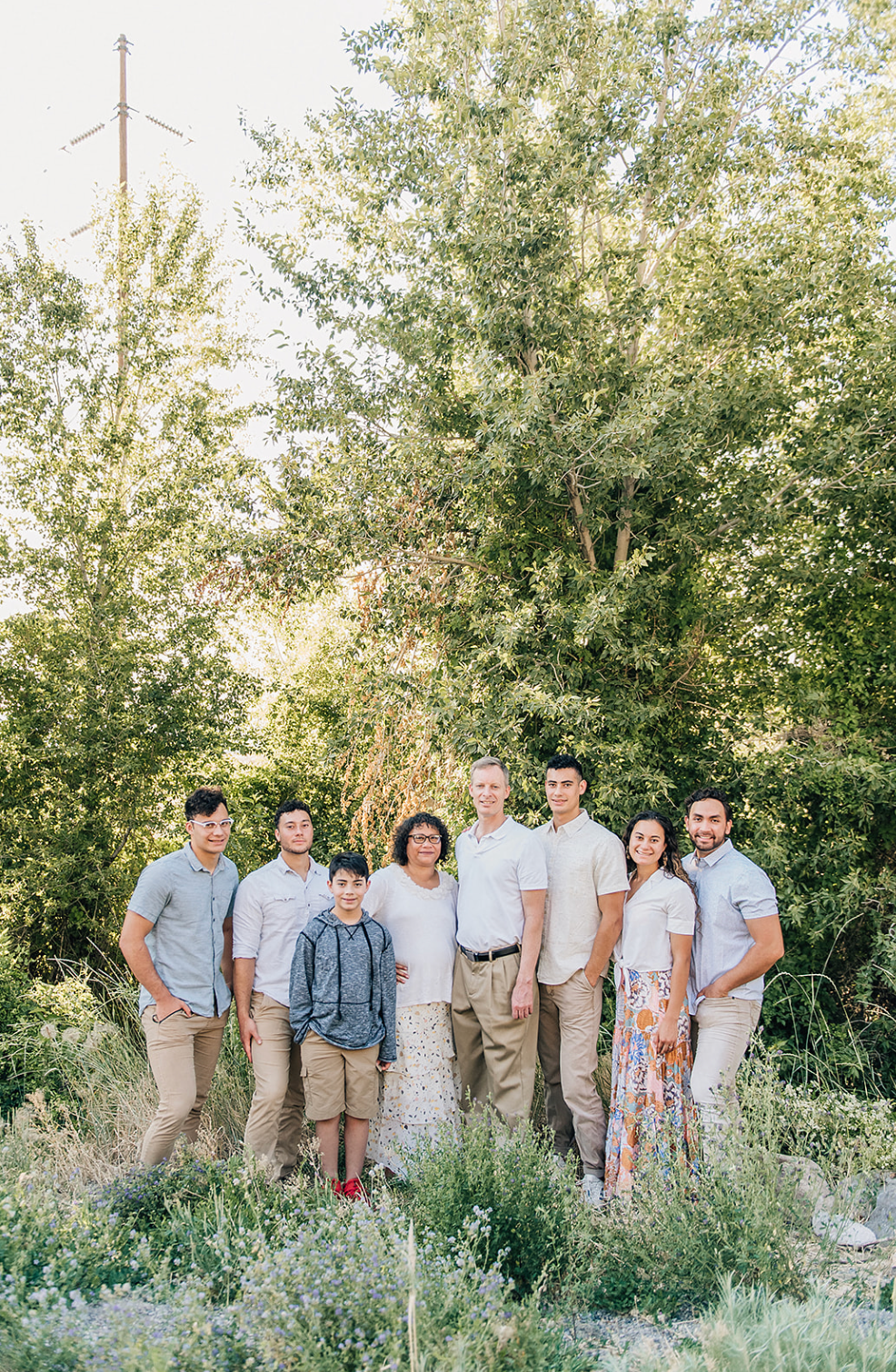 Group of people in coordinated light outfits standing together outdoors against a backdrop of green trees.