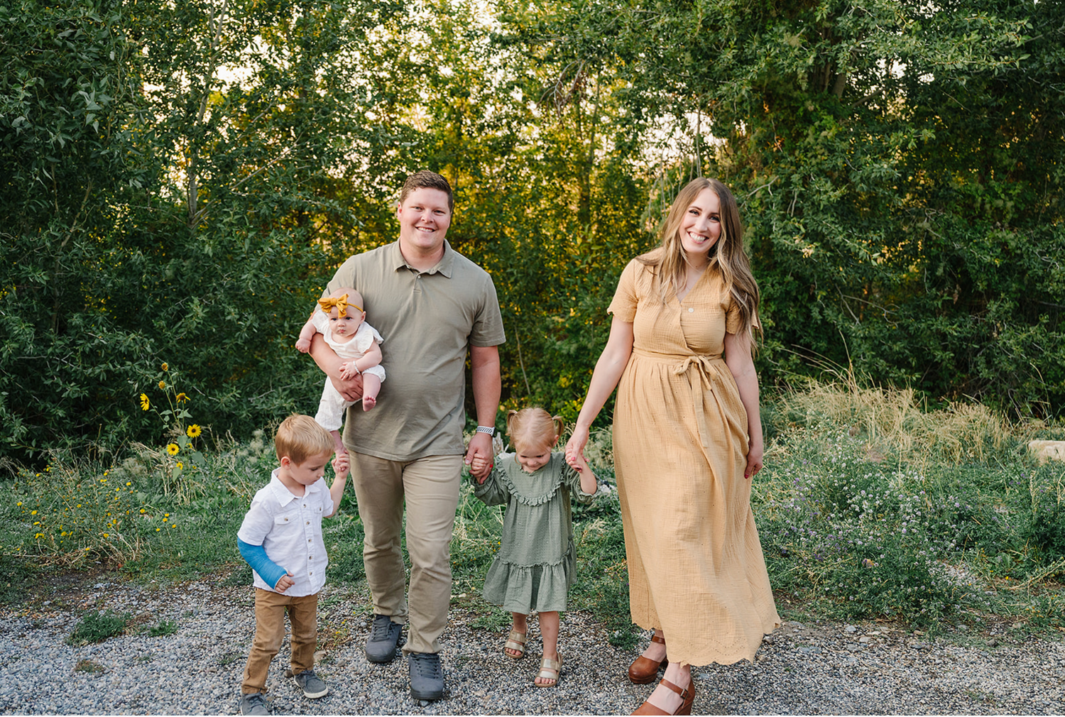 A family wearing coordinated neutral outfits walks together on a gravel path surrounded by greenery at sunset.