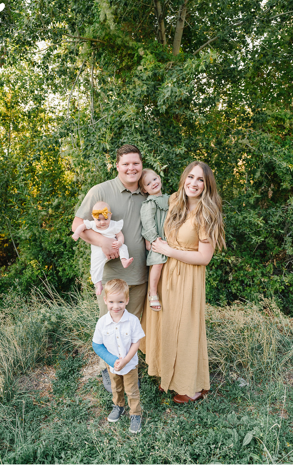 A family poses together outdoors against a backdrop of lush green foliage on a sunny summer day.