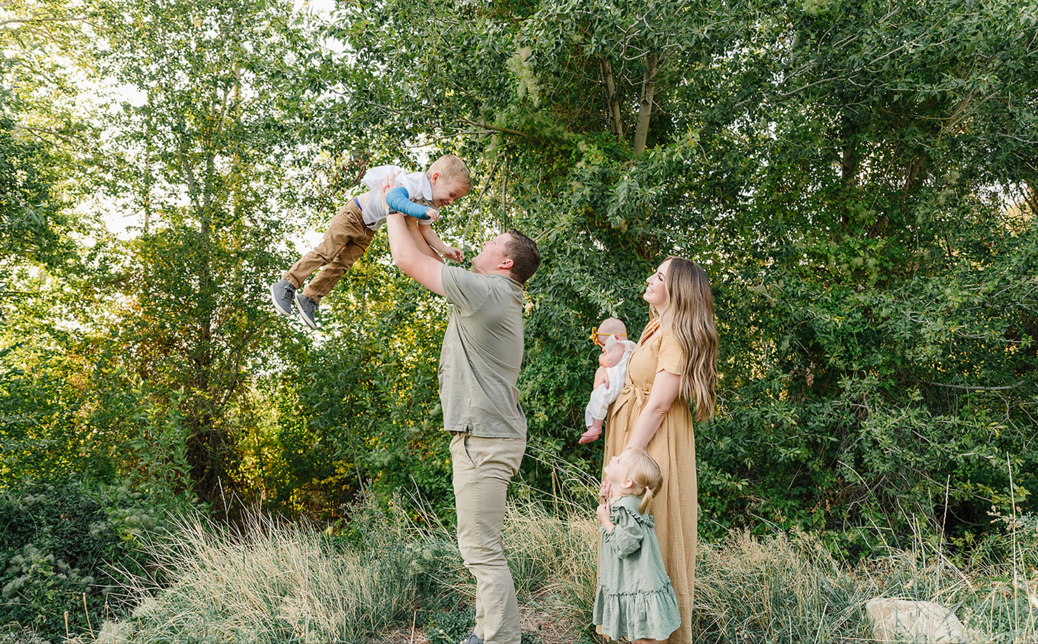 A family enjoys a tender outdoor moment together in a sunlit natural setting among green trees and grass.