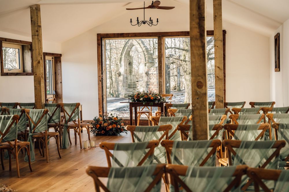 Spacious wedding reception hall with wooden beams, cross-back chairs and large windows.