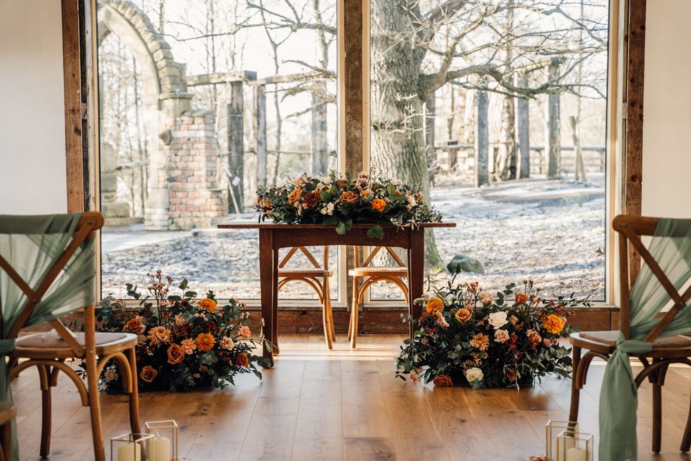 Rustic wedding ceremony setup with orange floral arrangements and wooden chairs in a bright windowed venue.