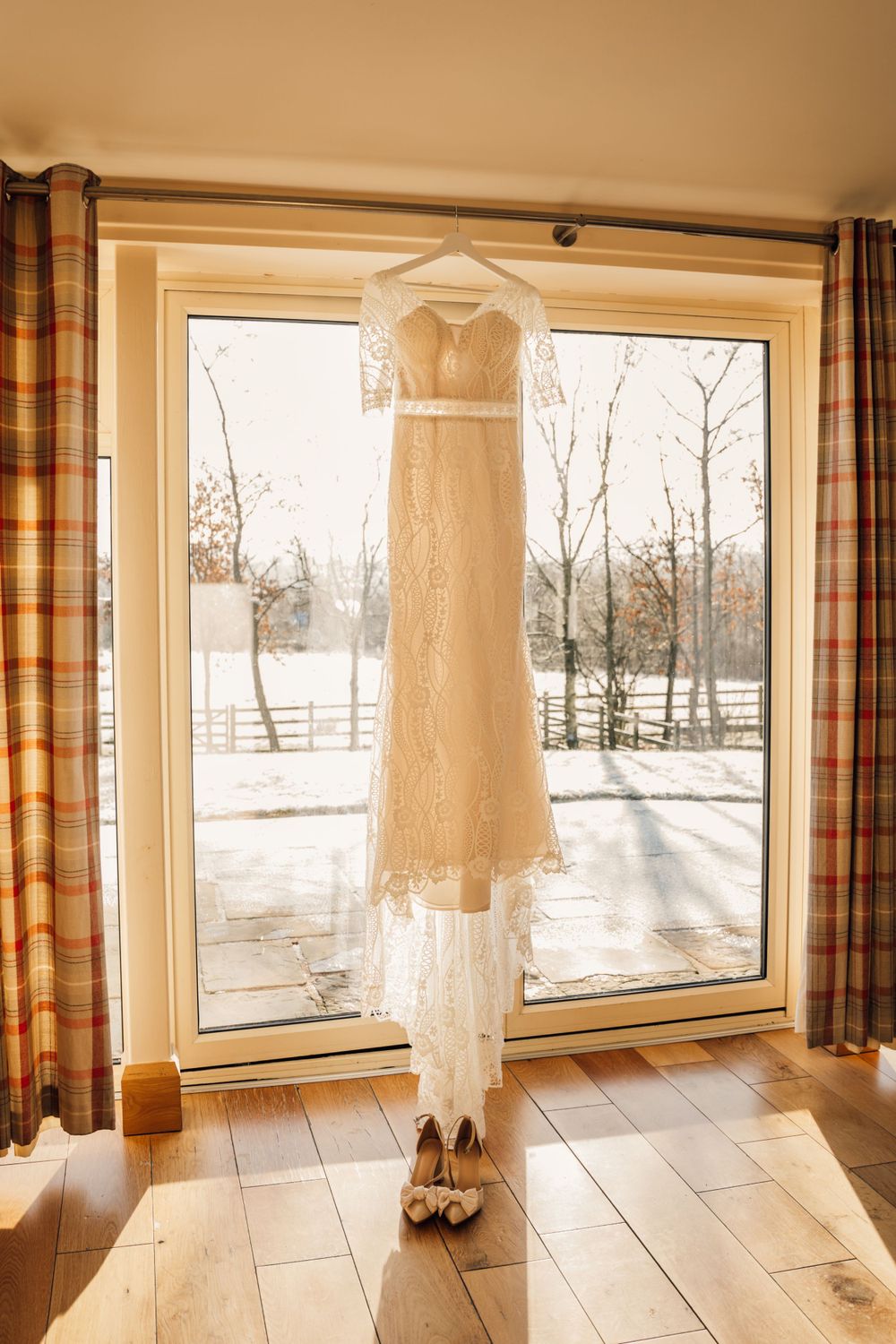 White lace wedding dress hanging against window with shoes displayed on wooden floor.