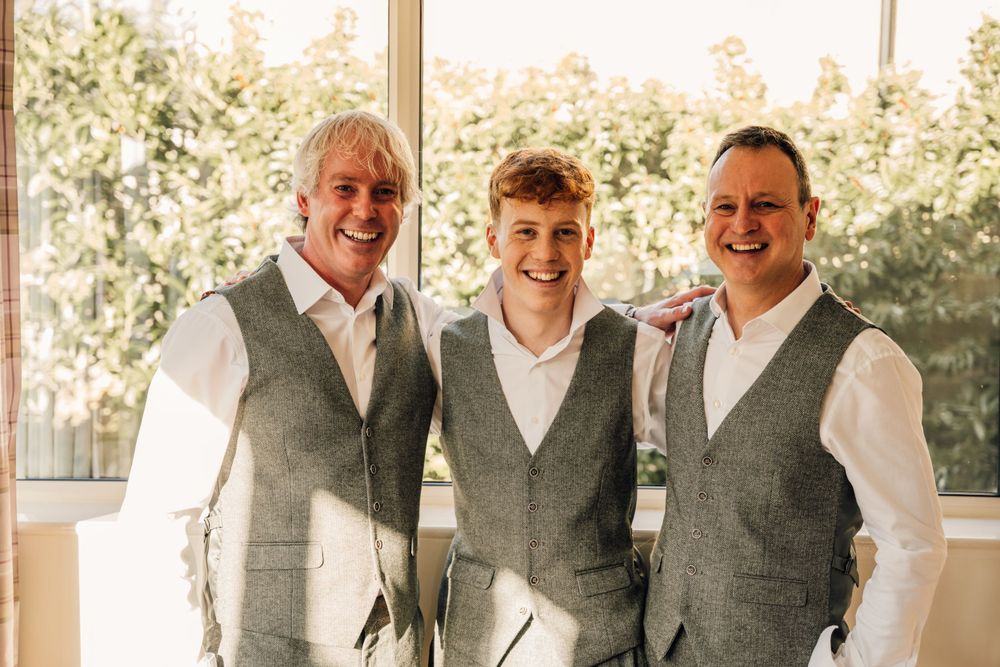 Wedding party members in matching grey vests and white shirts pose by window.