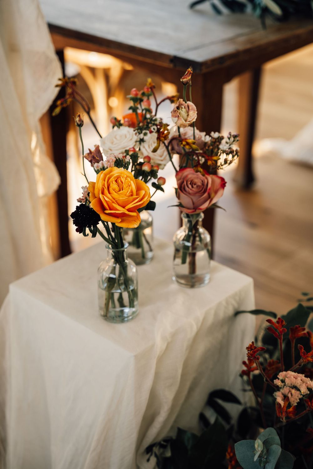 Mason jar centerpieces with orange and pink roses on white tablecloths in rustic wedding decor.