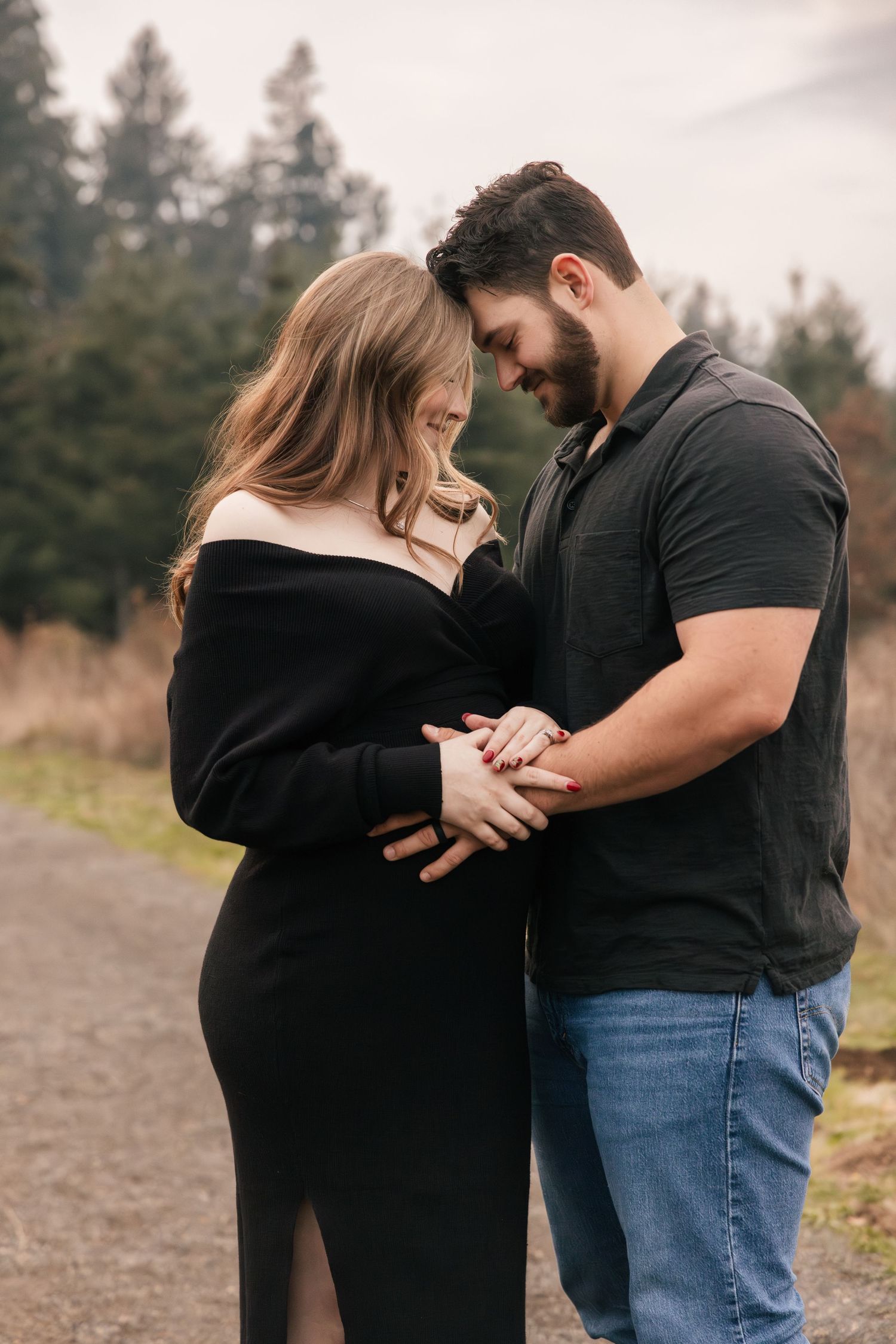 A couple shares an intimate moment on a dirt path surrounded by evergreen trees on an overcast day.