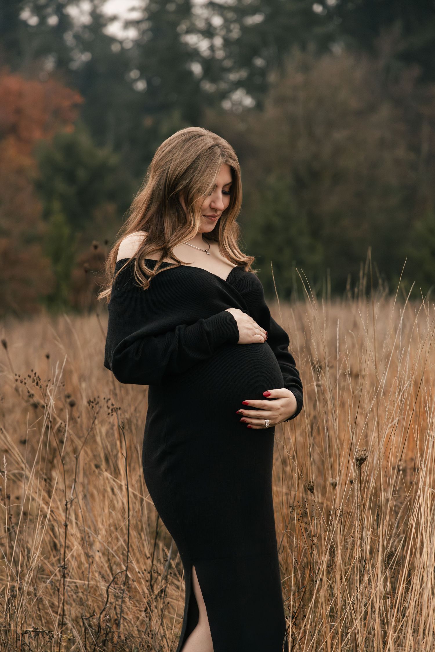 A person in a black dress cradles their pregnant belly while standing in tall autumn grass during a maternity photoshoot.