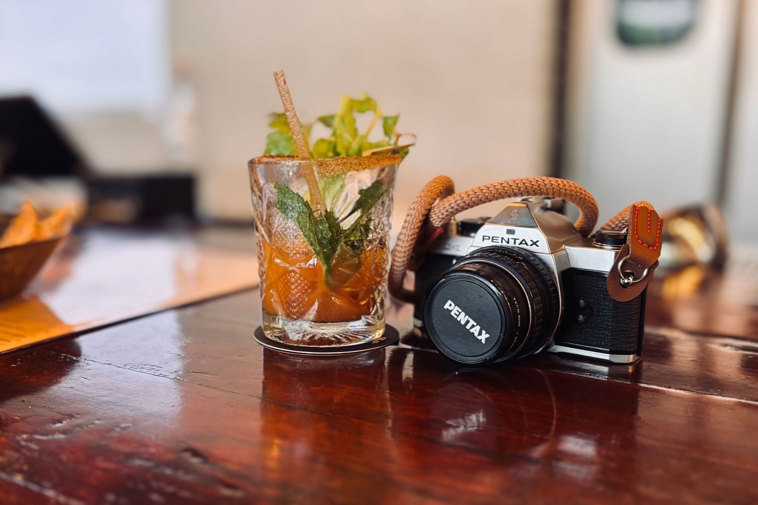 A vintage camera with brown leather strap sits beside a crafted cocktail garnished with herbs and citrus on a wooden table.