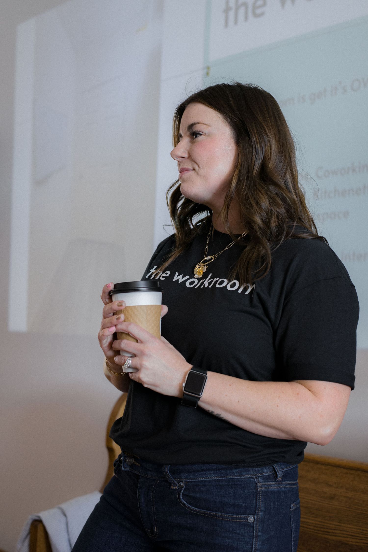 Woman in a black The Workroom t-shirt holding a coffee cup while presenting in front of a projector.