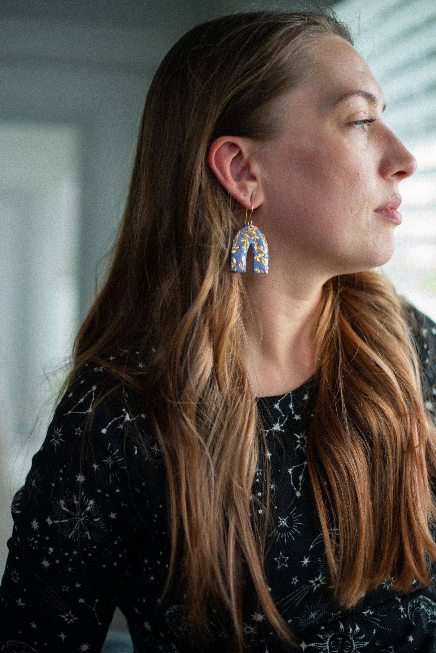 Woman with long auburn hair wearing blue arch earrings and a constellation-print black top gazing away.