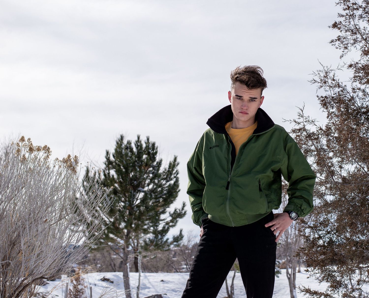 Teen male model in green jacket stands among snow-covered trees in winter.