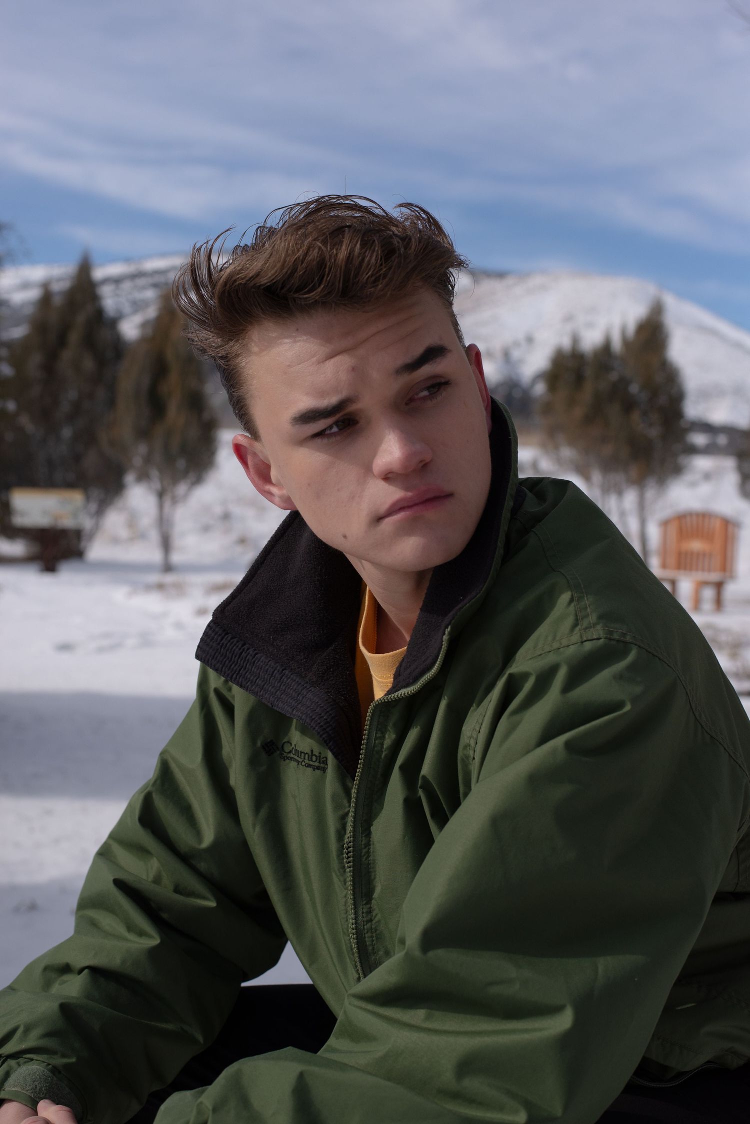 Young man in green Columbia jacket poses outdoors in snowy mountain landscape.