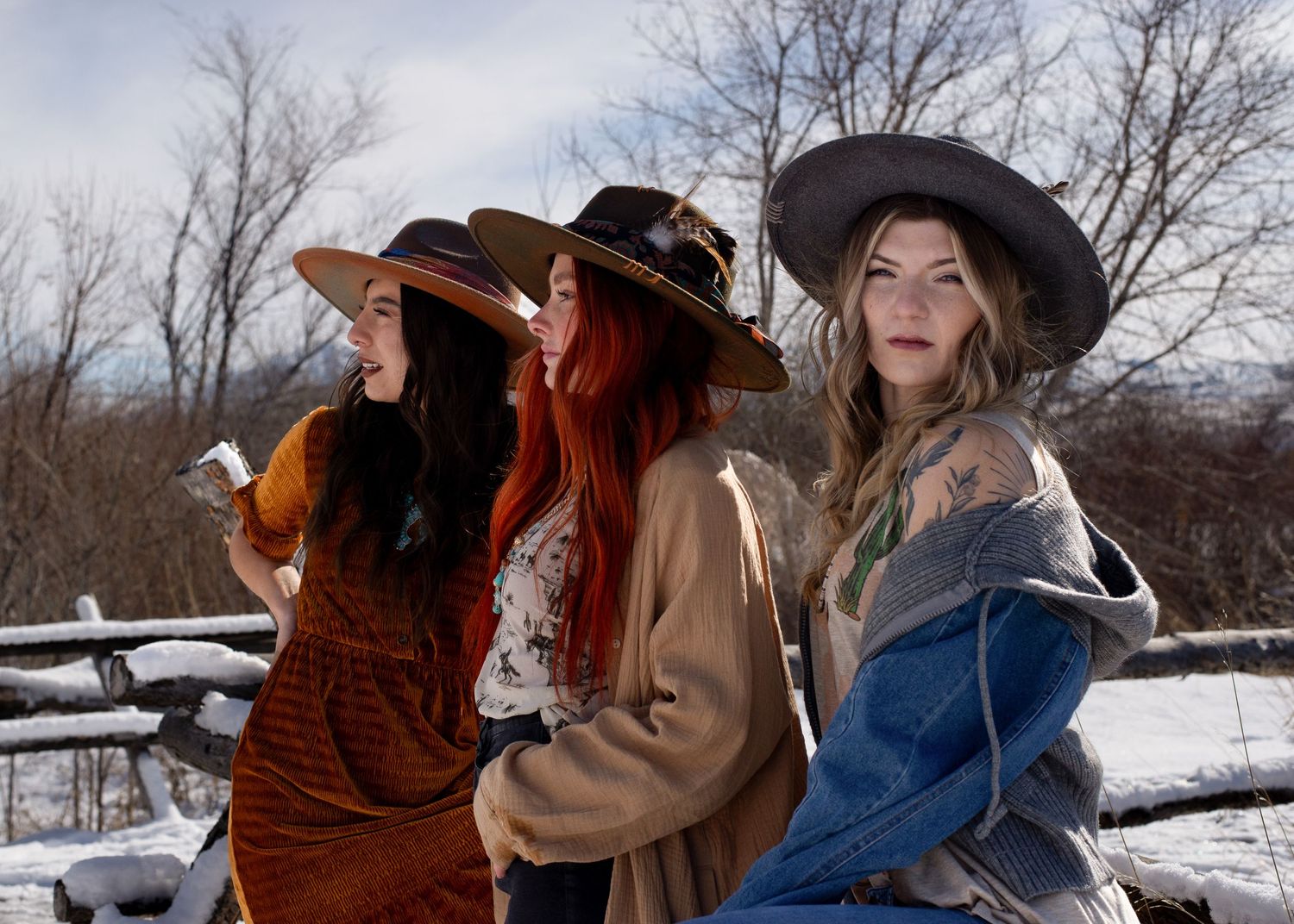 Three women in boho hats pose together at a snowy wooden fence outdoors.