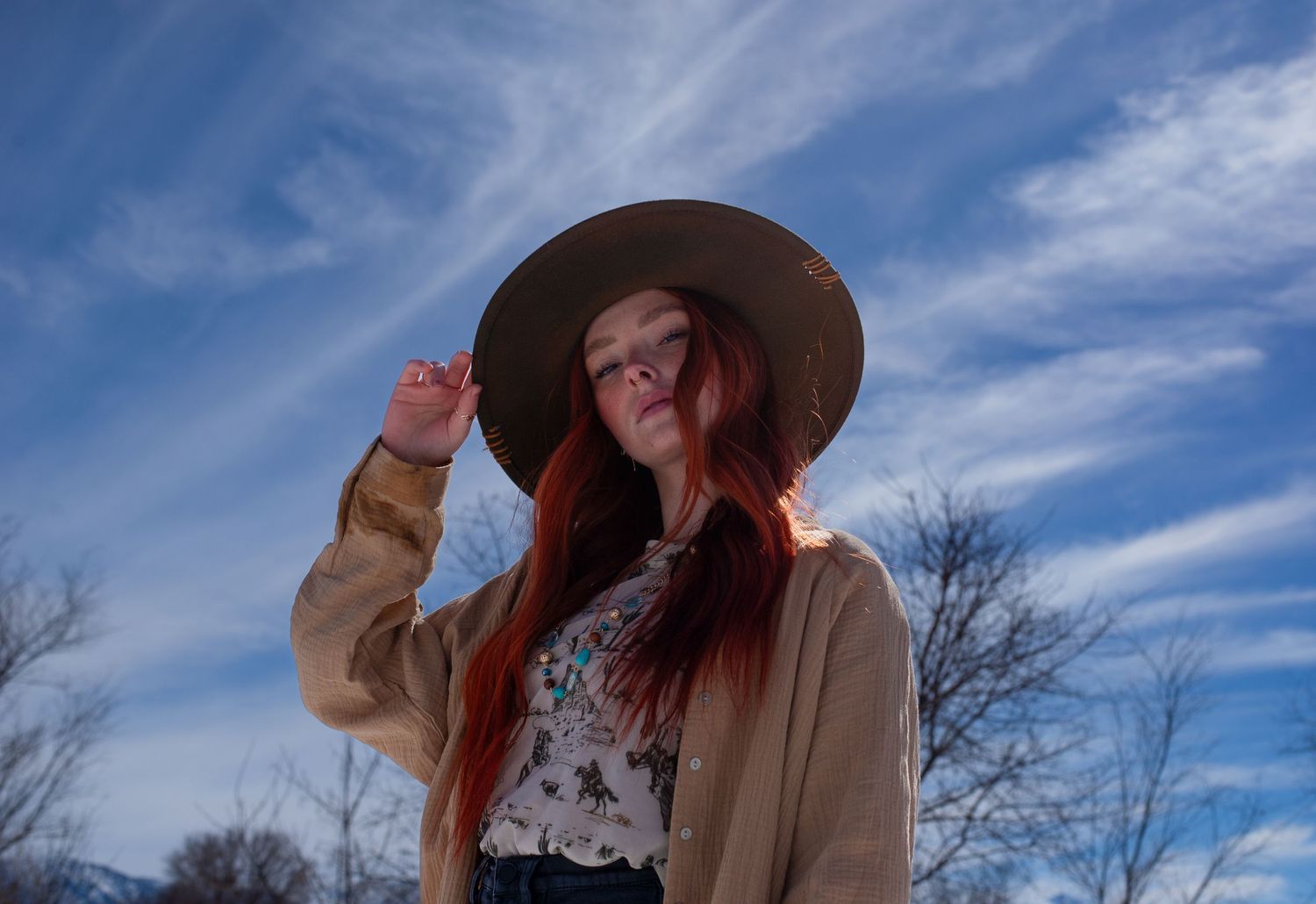 Red-haired woman in wide-brim hat poses under blue sky in winter setting.