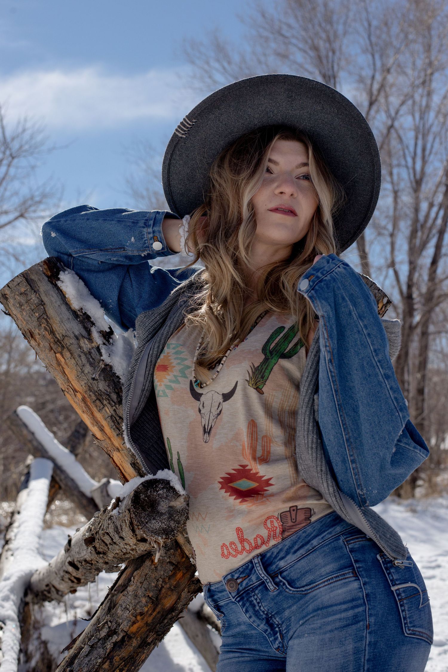 Woman in cowboy hat and Southwest print top leans on snowy log fence.