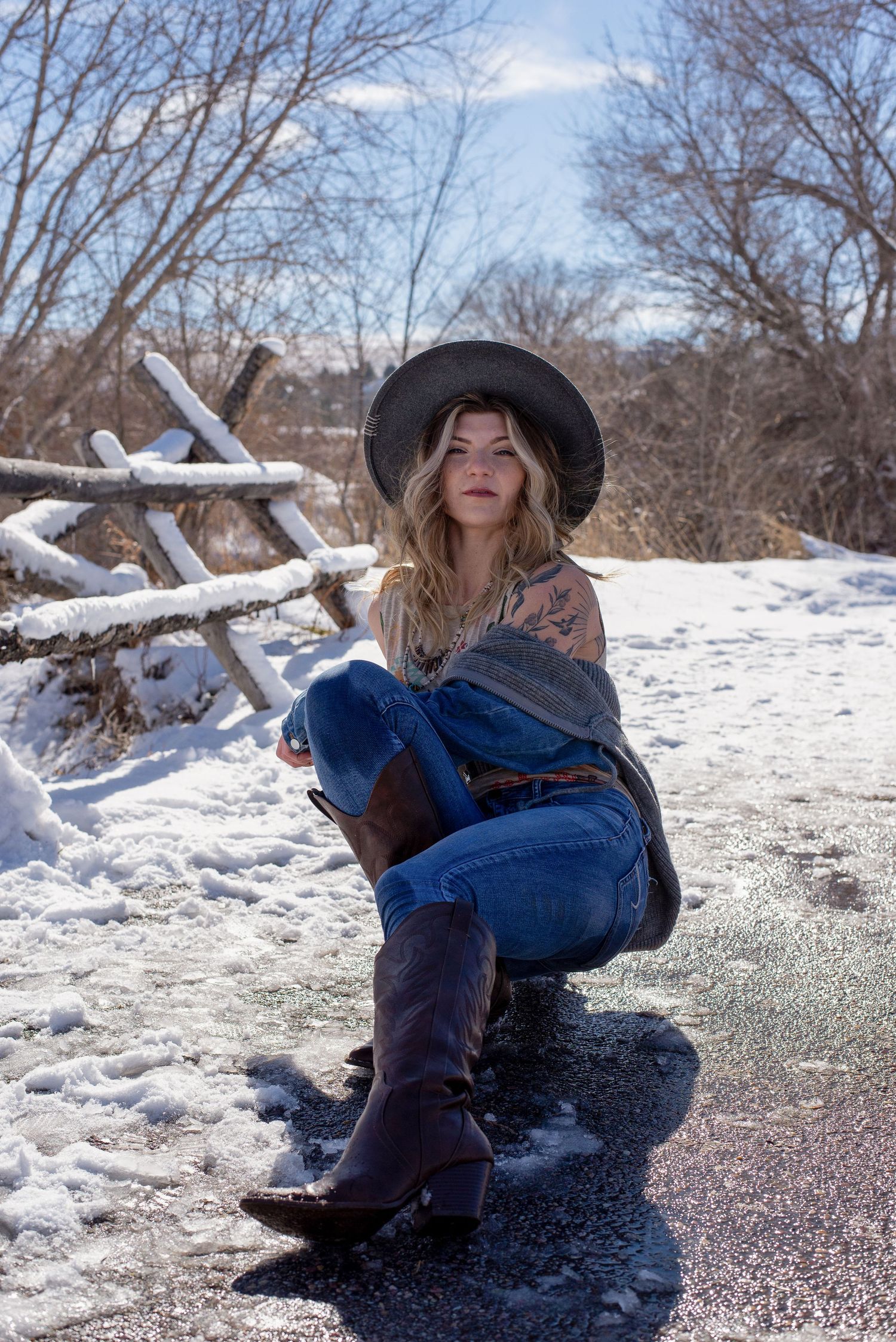 Blonde woman in cowboy hat and boots sits on snowy road in winter.