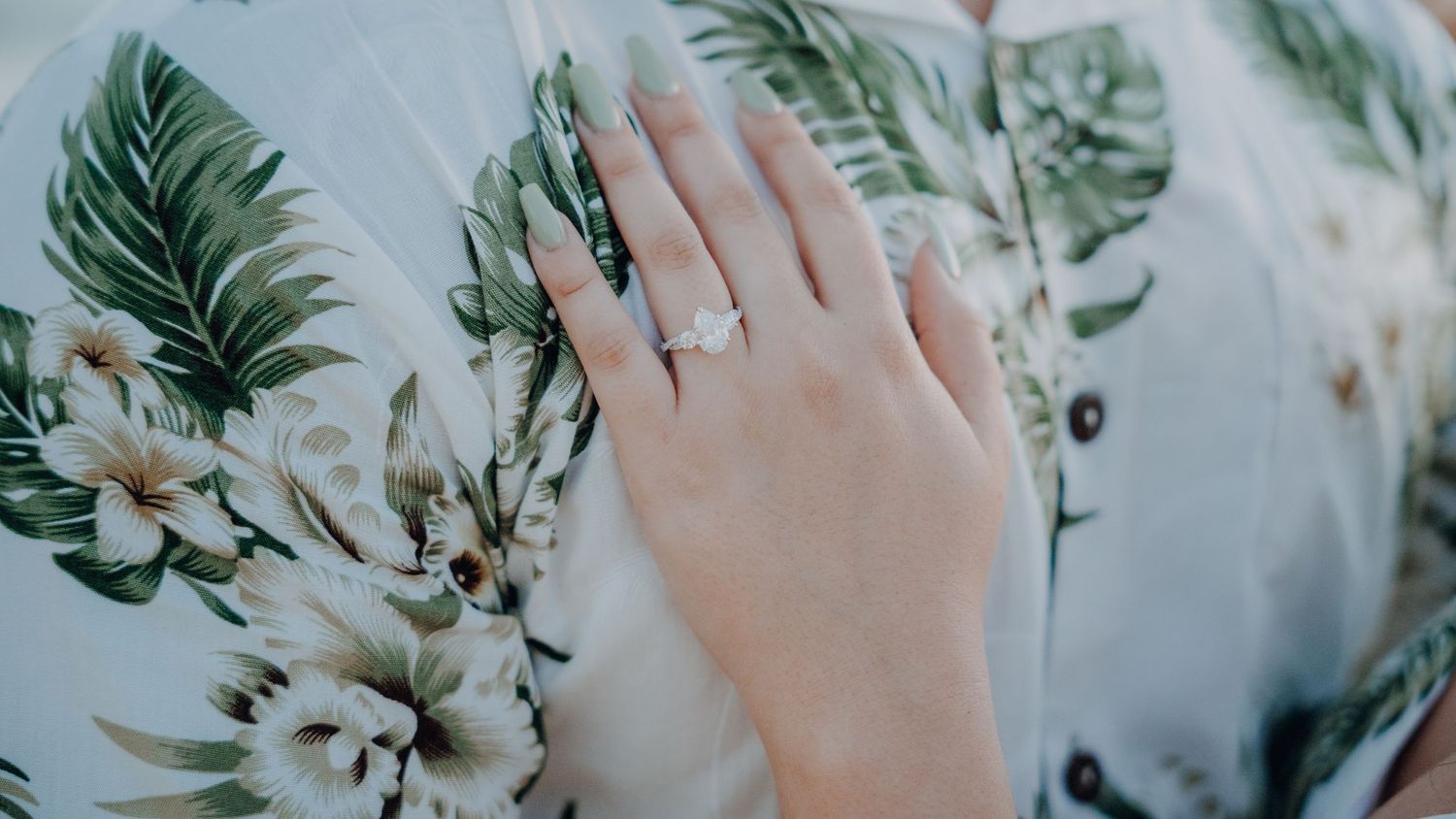 A hand adorned with an engagement ring set against tropical greenery and white flowers.