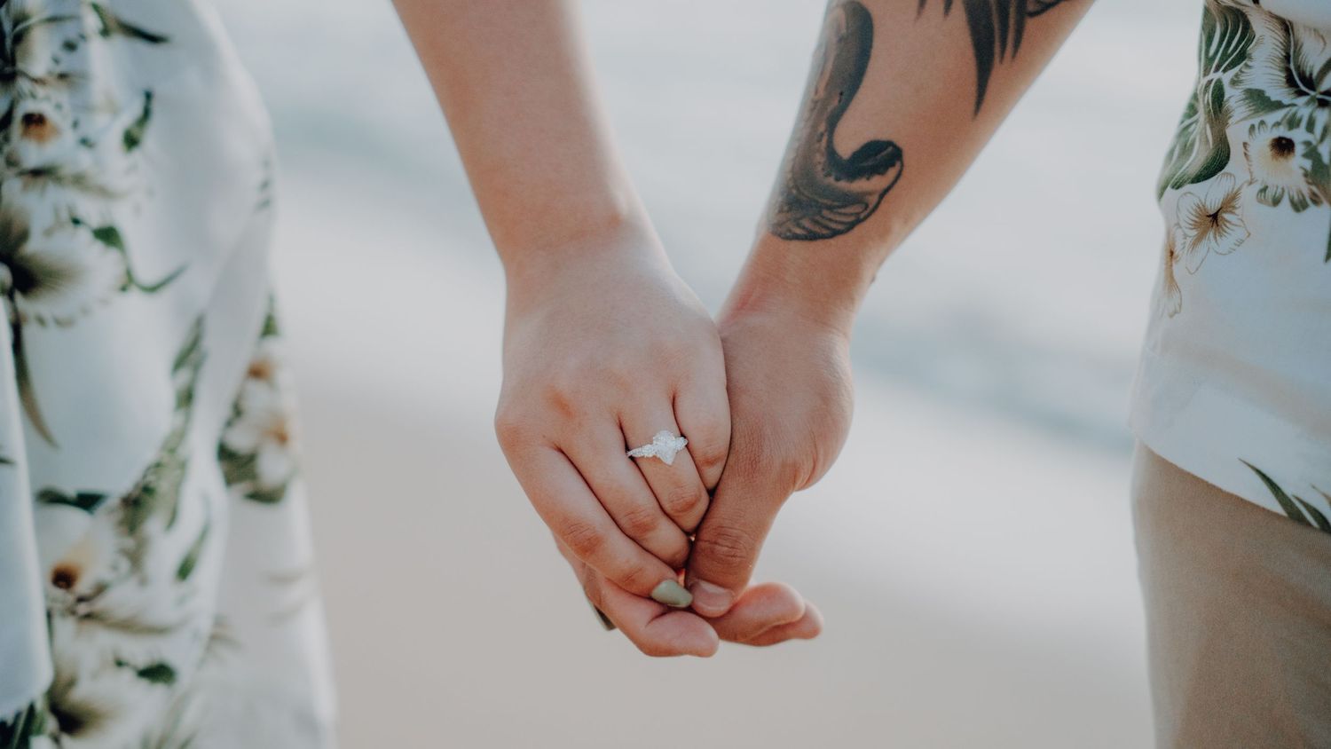 Two hands clasped together wearing matching rings with green foliage in the background.