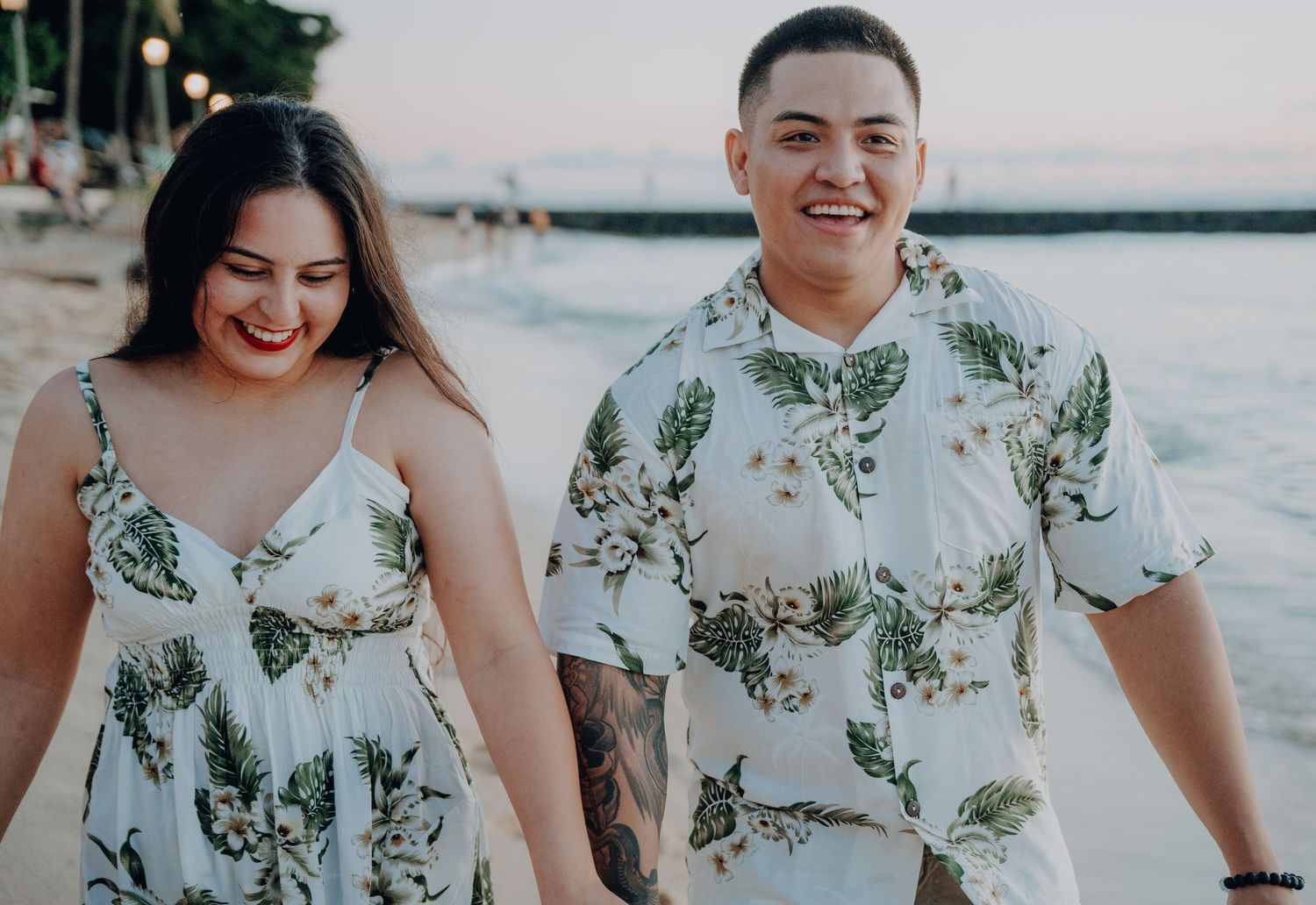 A couple wearing coordinating tropical print outfits smile while walking on the beach.