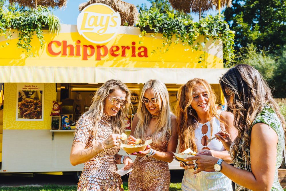 A group of friends enjoying food and drinks together outside a yellow Chipperia restaurant on a sunny day.