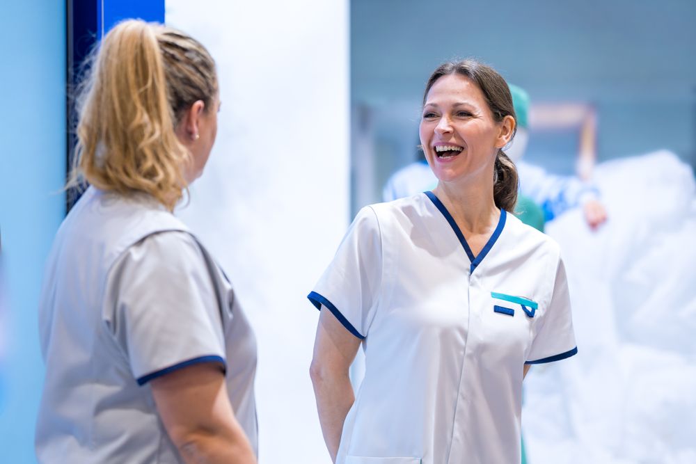 Two healthcare workers in white and blue nursing uniforms share a cheerful moment during work at a medical facility.