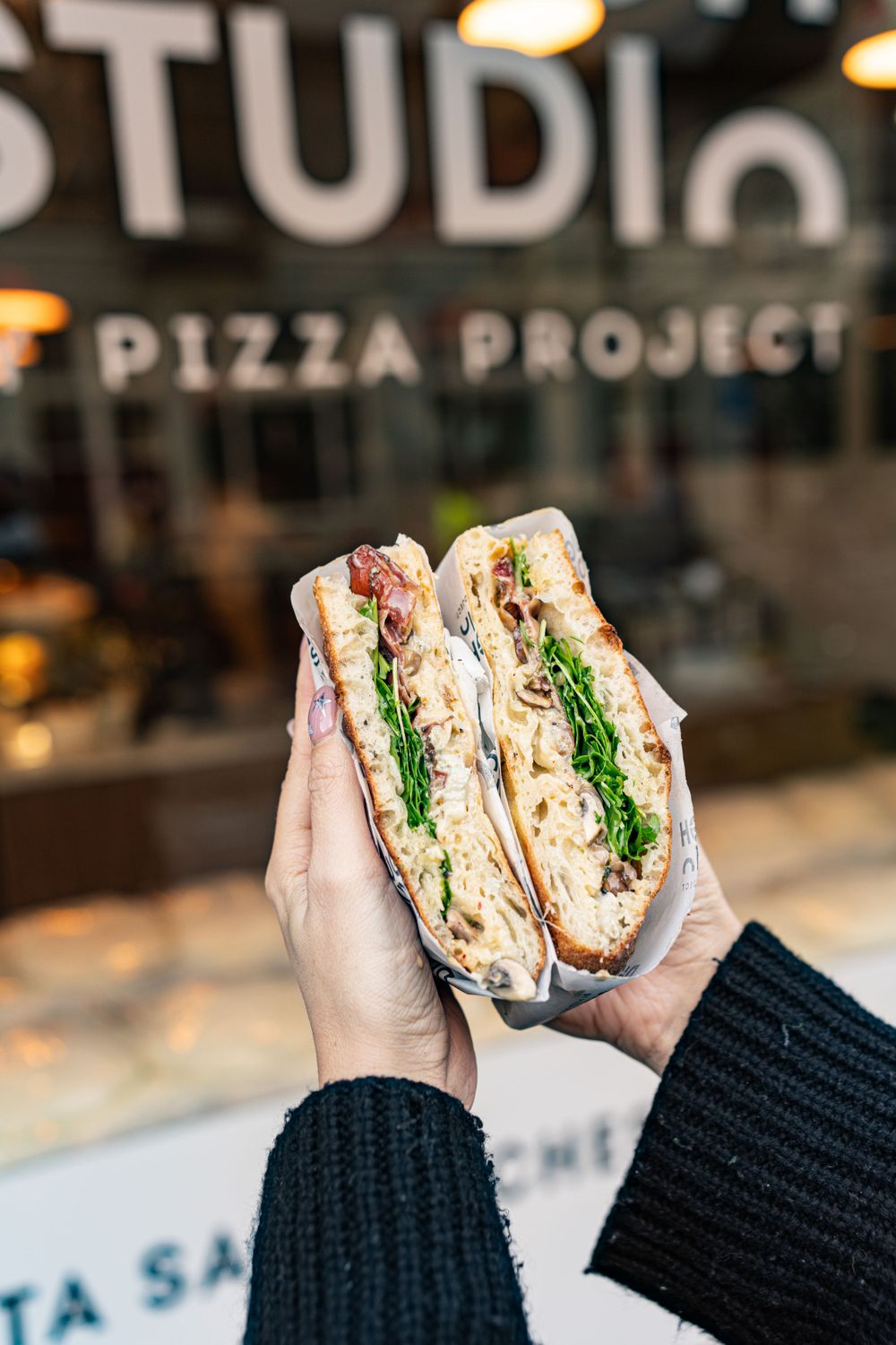 Hands holding a fresh sandwich with turkey, greens and vegetables inside a cafe with Studio Pizza Project signage.
