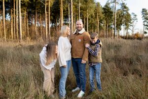 Family of four poses together in tall grass with pine trees in background.