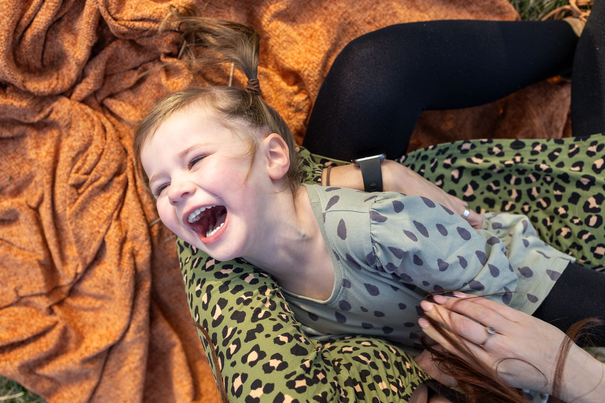 Child laughing joyfully while laying on leopard print and orange blankets outdoors.