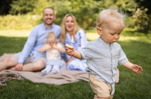 Toddler explores grassy area while family relaxes on picnic blanket in background.