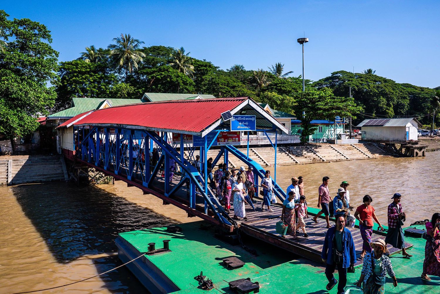 Tourists gather on a colorful dock with red roof and blue railings at a tropical waterfront location.