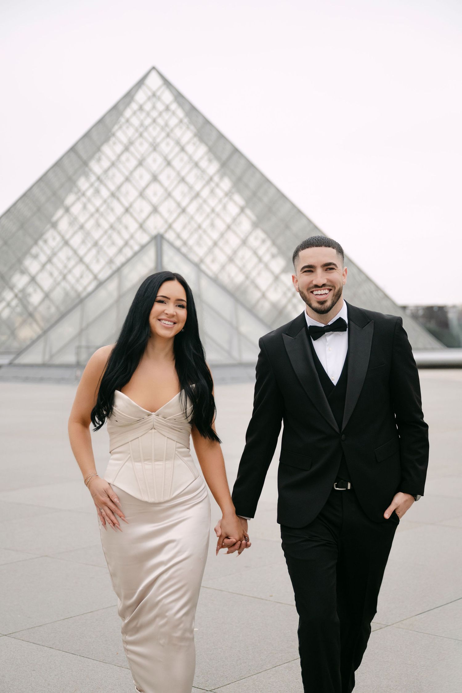 Elegant couple in formal attire poses in front of the iconic glass pyramid at the Louvre Museum in Paris.