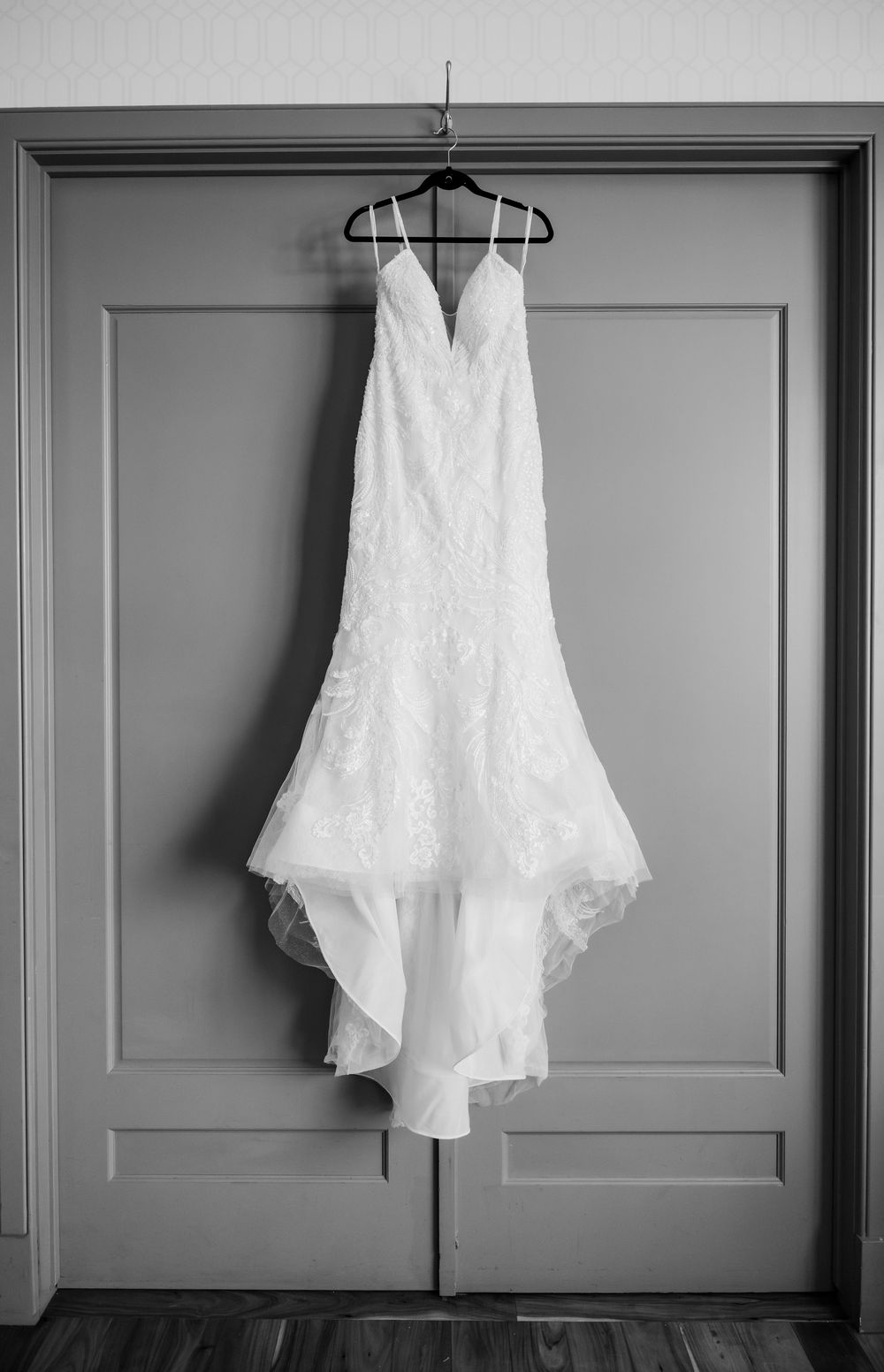 A white lace wedding dress hangs elegantly against a gray paneled wall in a monochromatic setting.