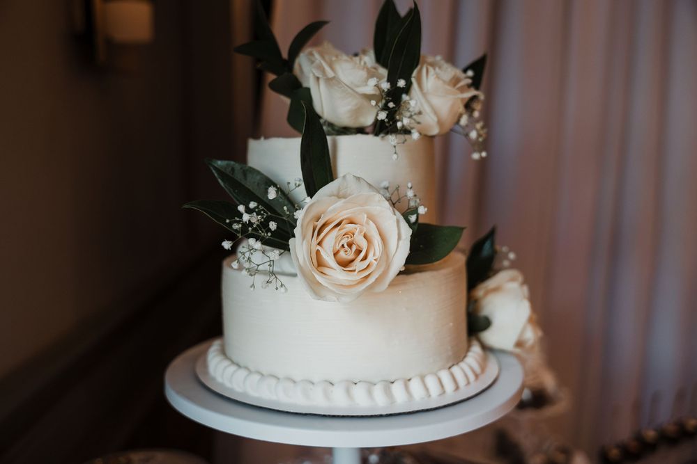 A white tiered wedding cake decorated with fresh pink roses and eucalyptus leaves sits elegantly on display.