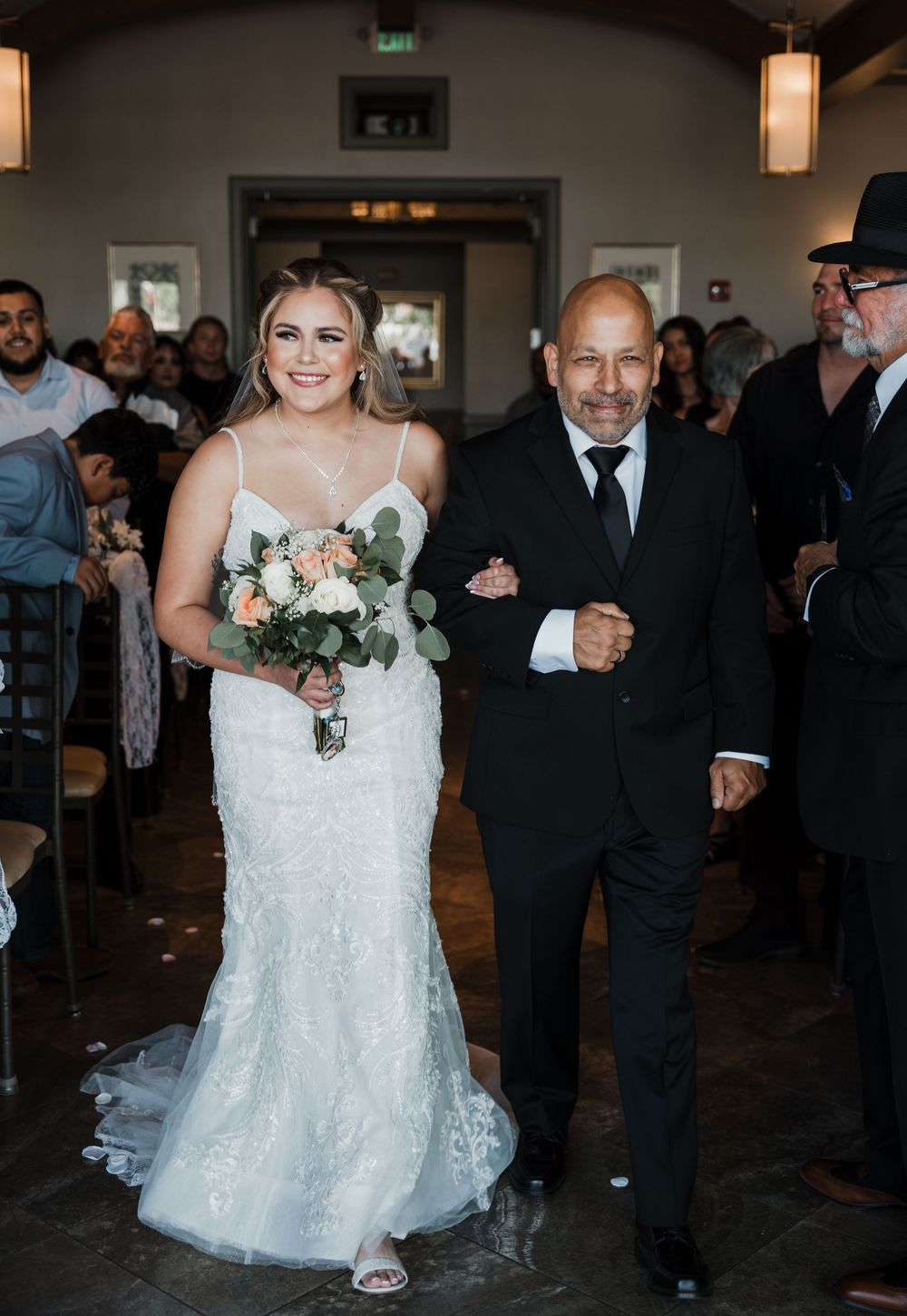 Wedding ceremony procession showing a bride in a white lace dress walking down the aisle at an indoor venue.