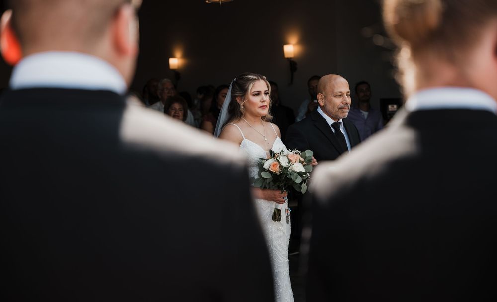 A bride in a white dress walks down the aisle with her escort during an indoor evening wedding ceremony.