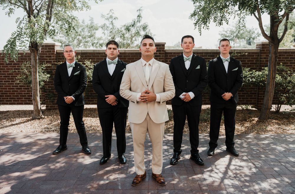 A wedding party in formal attire poses together under trees on a sunny day near a brick wall.