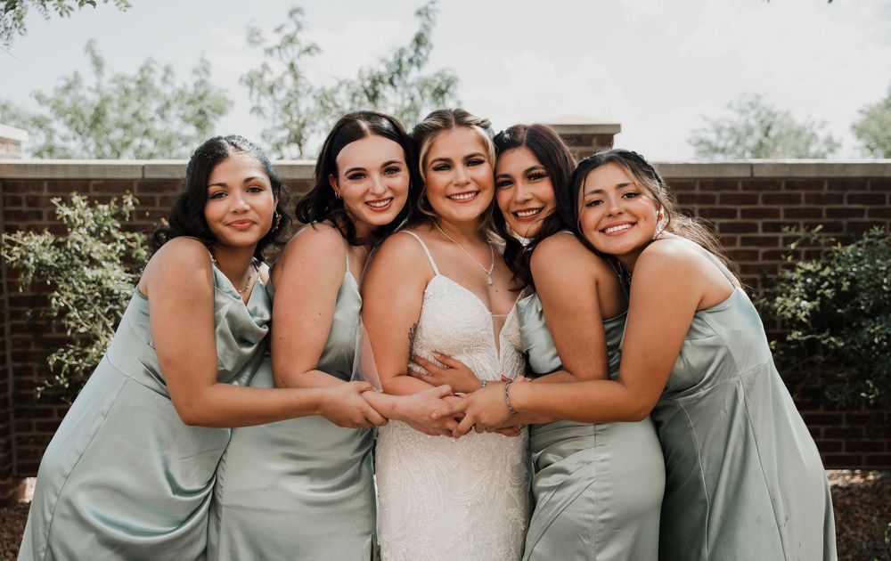 Bridesmaids in sage green dresses pose together with the bride in white for an outdoor wedding photo.