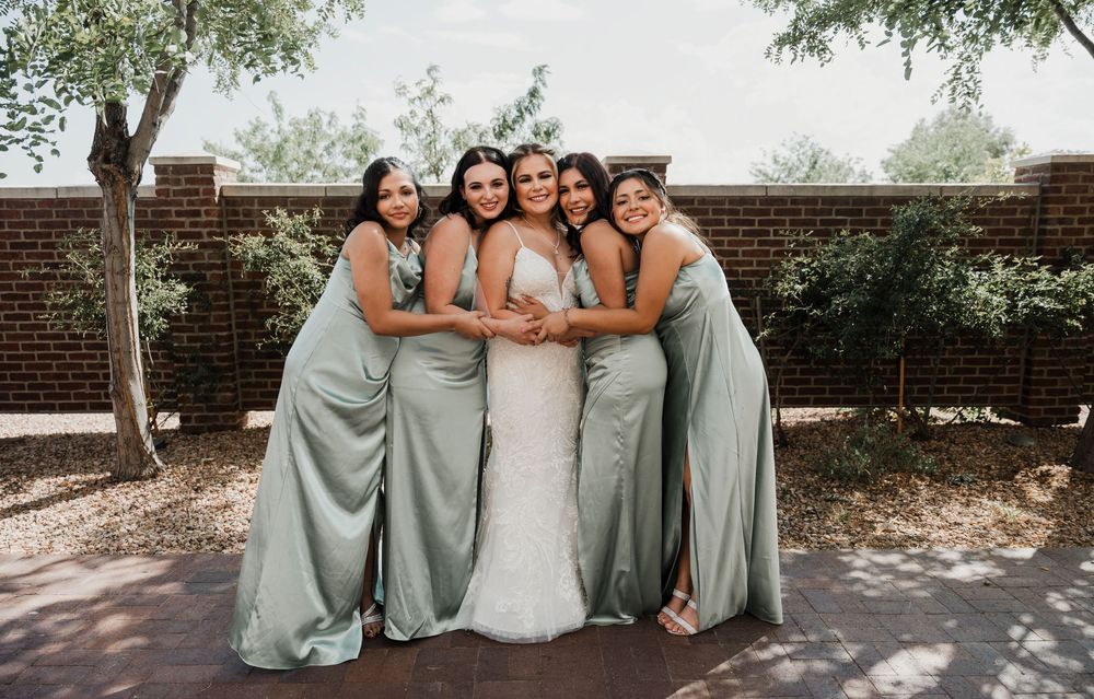 Wedding party in sage green dresses and white gown pose together outdoors near a brick wall.