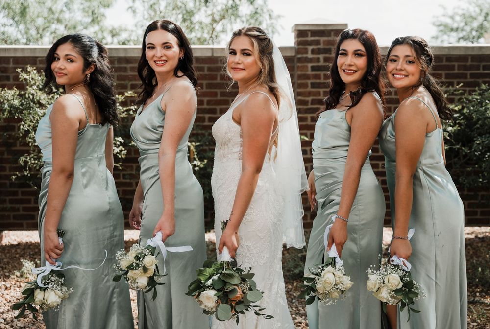 Bridesmaids wearing sage green dresses stand in a line outdoors for wedding photos.